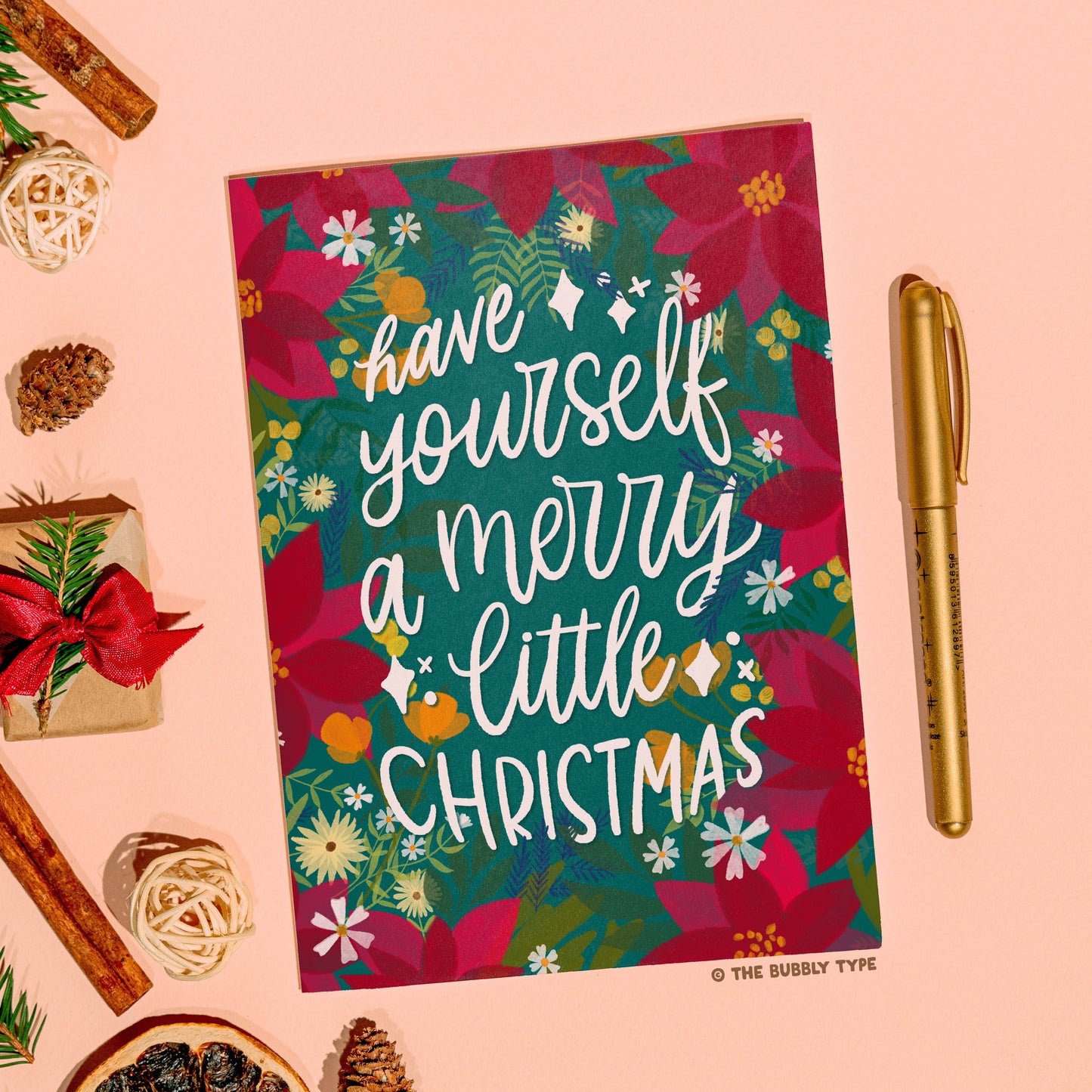 The same Christmas card displayed on a peach background. To the left are festive trinkets including baubles and greenery, while a gold pen is placed on the right side of the card. Australian made Christmas cards. Have yourself a merry littler christmas, hand drawn design. Affordable Christmas gift card