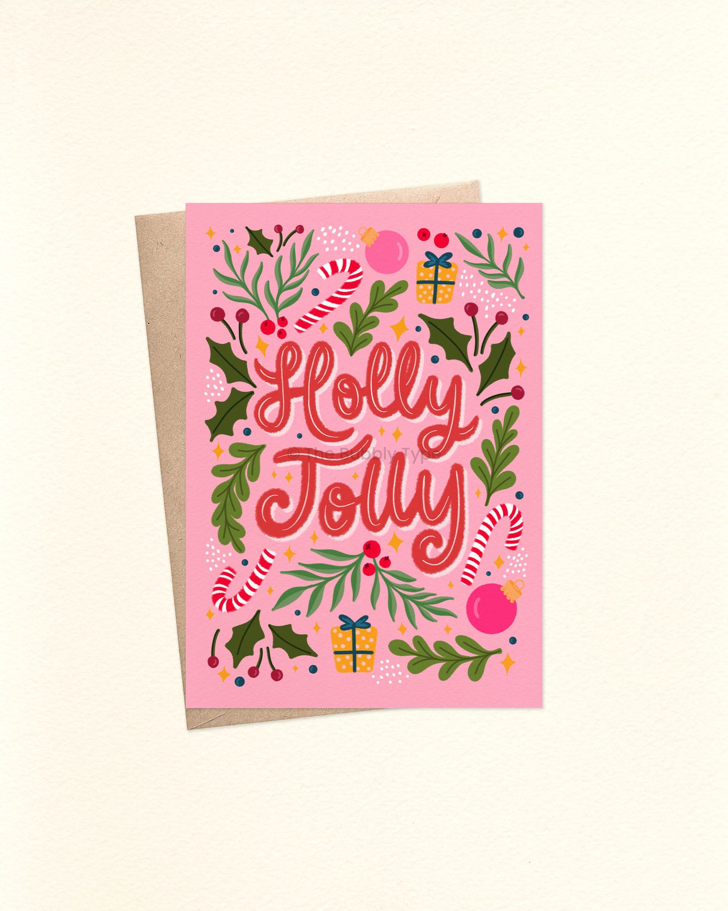 A6 Christmas card with a pale pink background and bold red hand-lettered text reading “Holly Jolly” in the centre. Surrounding the text are festive illustrations including holly leaves, red berries, candy canes, wrapped presents, baubles, and other foliage. The card is laid flat with a brown kraft envelope beneath it.