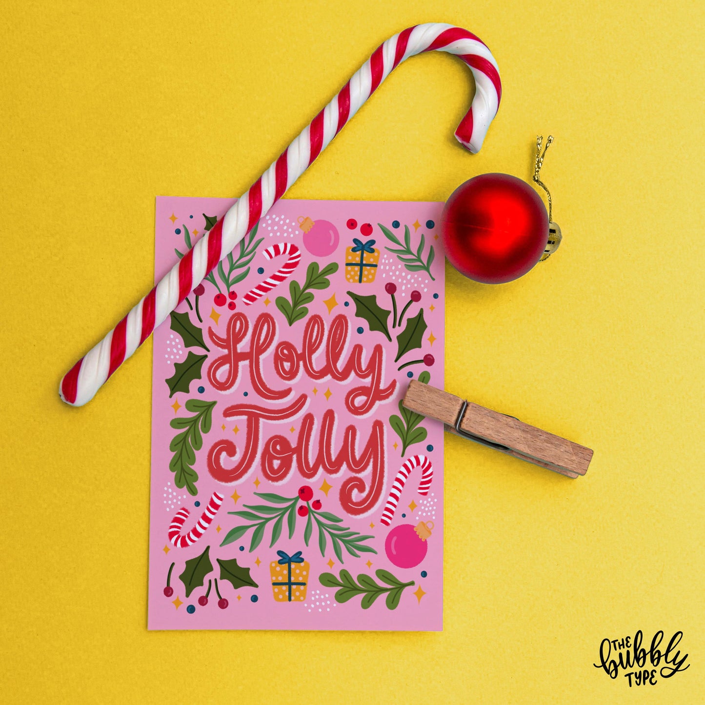 The “Holly Jolly” Christmas card displayed on a bright yellow background with a red bauble to the side and a striped candy cane positioned above it. Holly Jolly Christmas Card, Best pink greeting cards made in Australia.