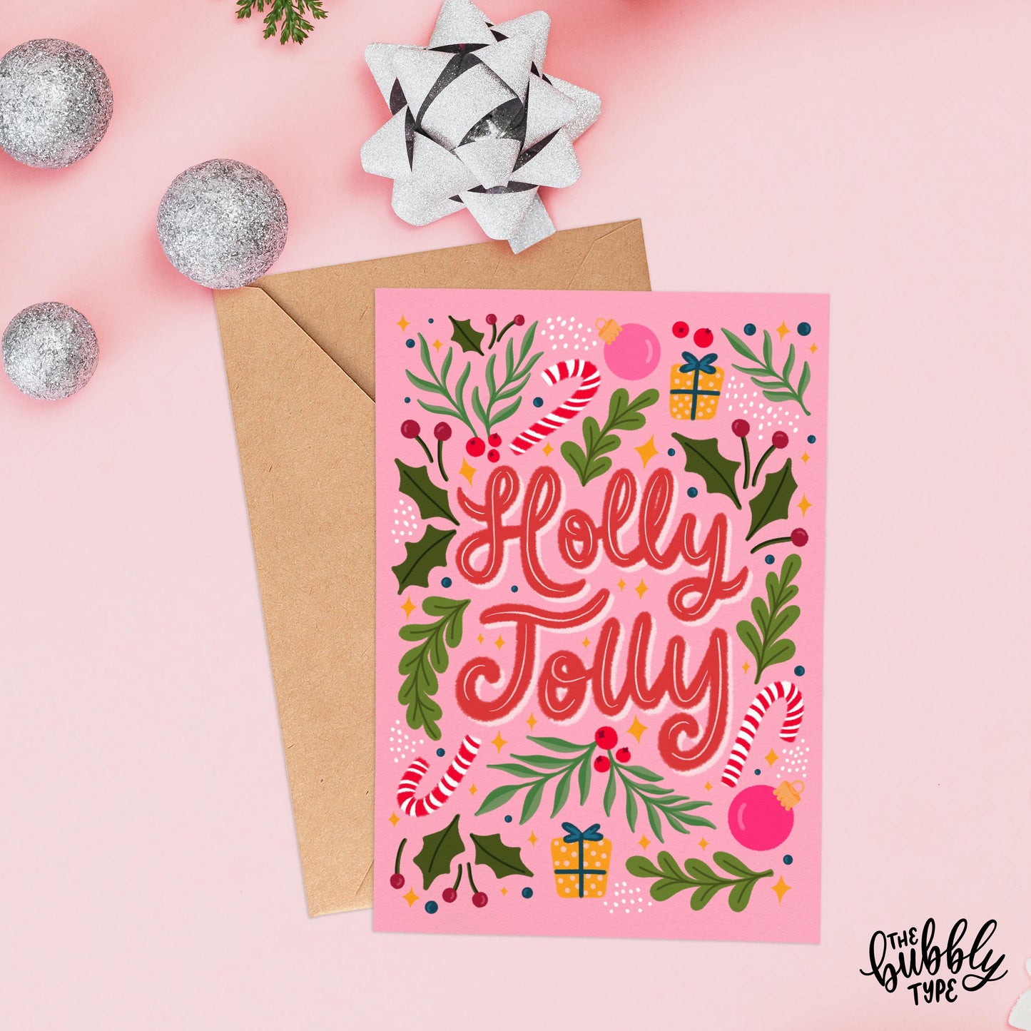 The same card laid flat on a light pink background alongside a kraft envelope. Decorative elements include silver baubles and a sparkly silver gift bow placed near the top of the image.
Holly Jolly greeting card, cute christmas card design, made in australia.