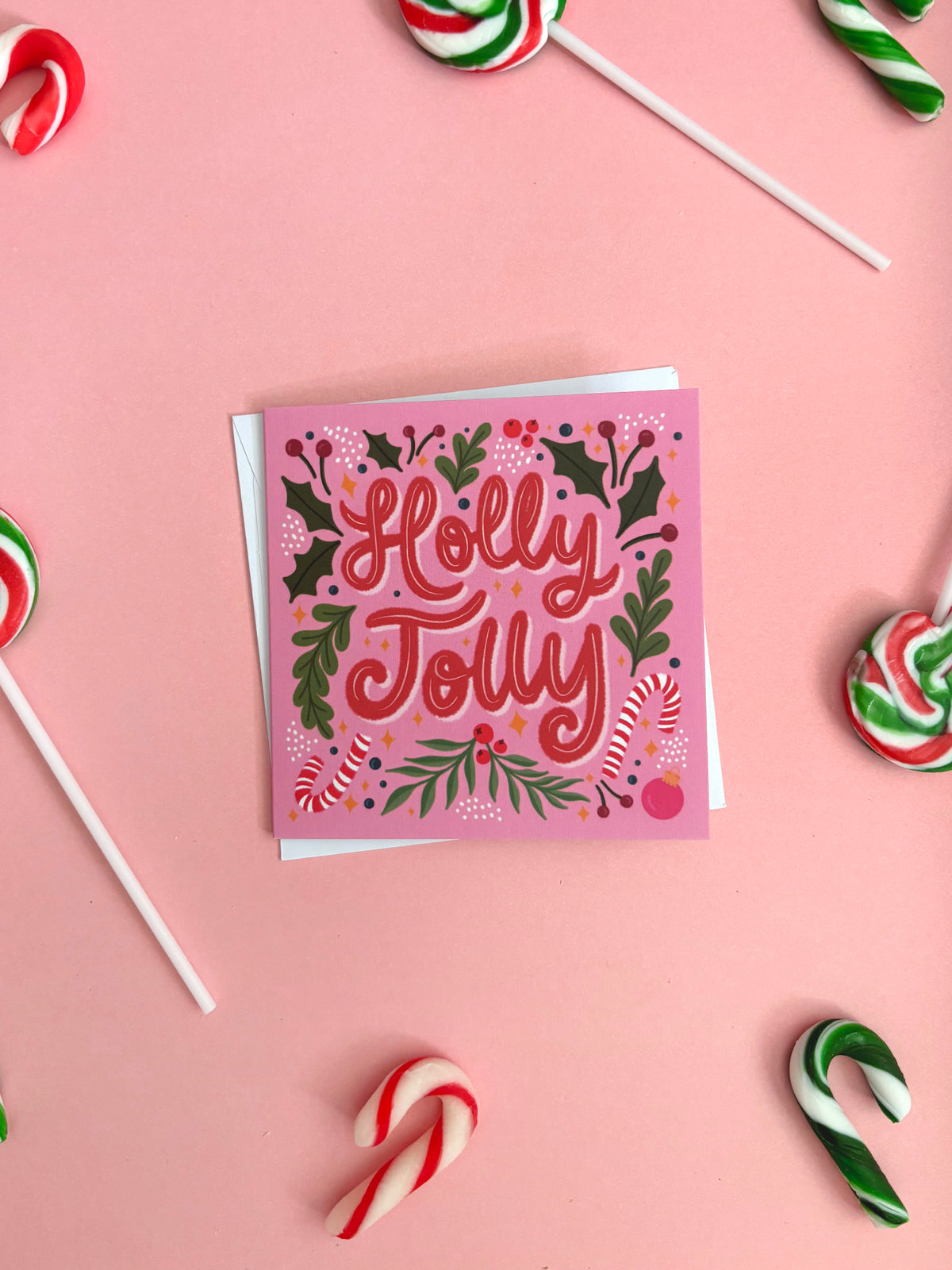 A square pink Christmas greeting card with 'Holly Jolly' written in red script, surrounded by holly leaves, berries, and candy canes, styled on a pink background with candy lollipops.