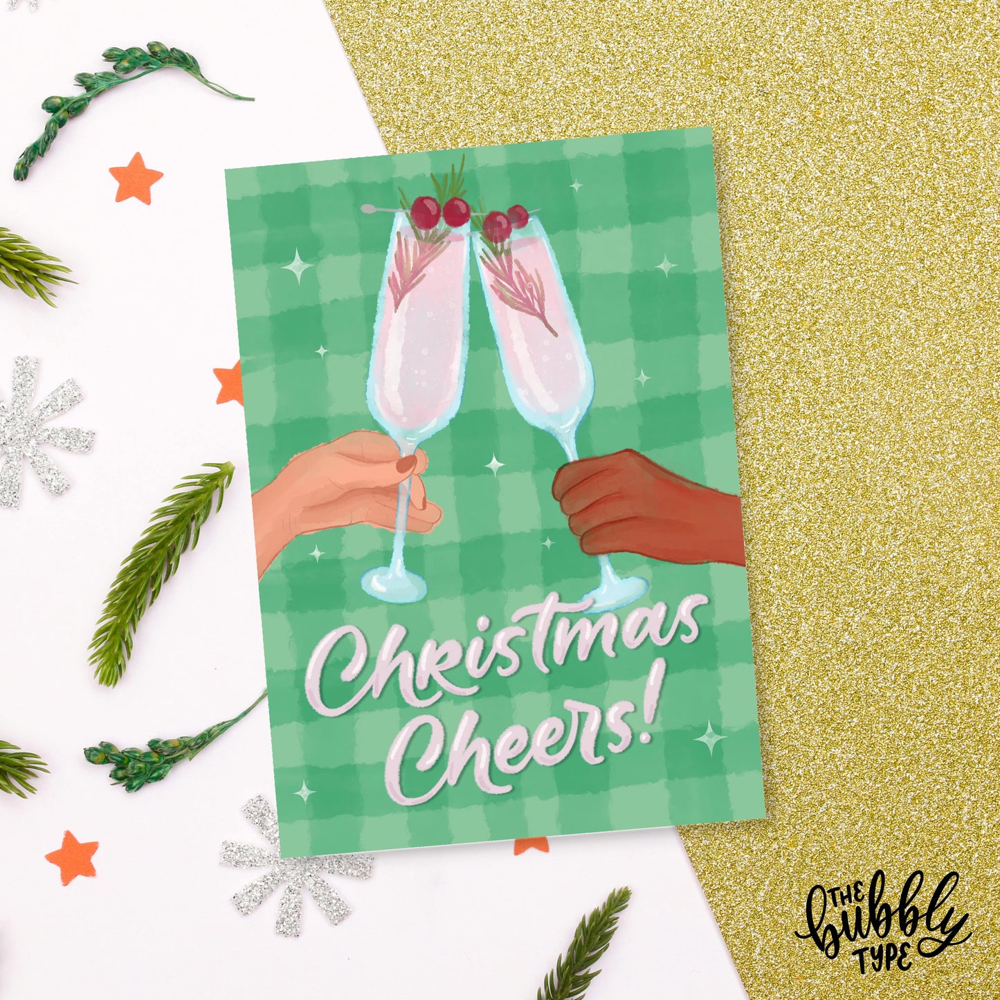 The front of the same A6 Christmas card placed flat on a background of white and gold star-patterned paper, surrounded by sprigs of pine leaves and silver glitter star decorations.
