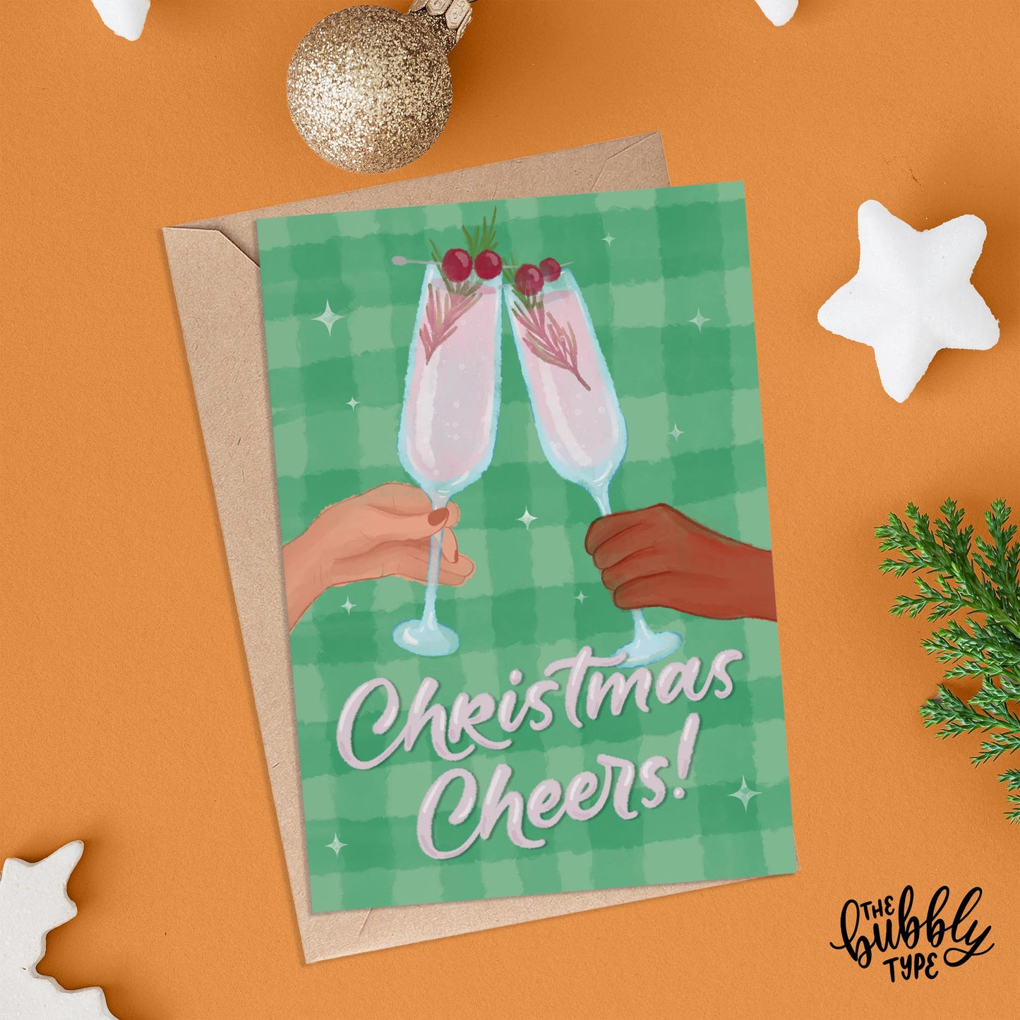Front of the “Christmas Cheers” card laid over a brown kraft envelope on an orange background. Around the card are festive decorations including white star ornaments and glittery gold baubles.