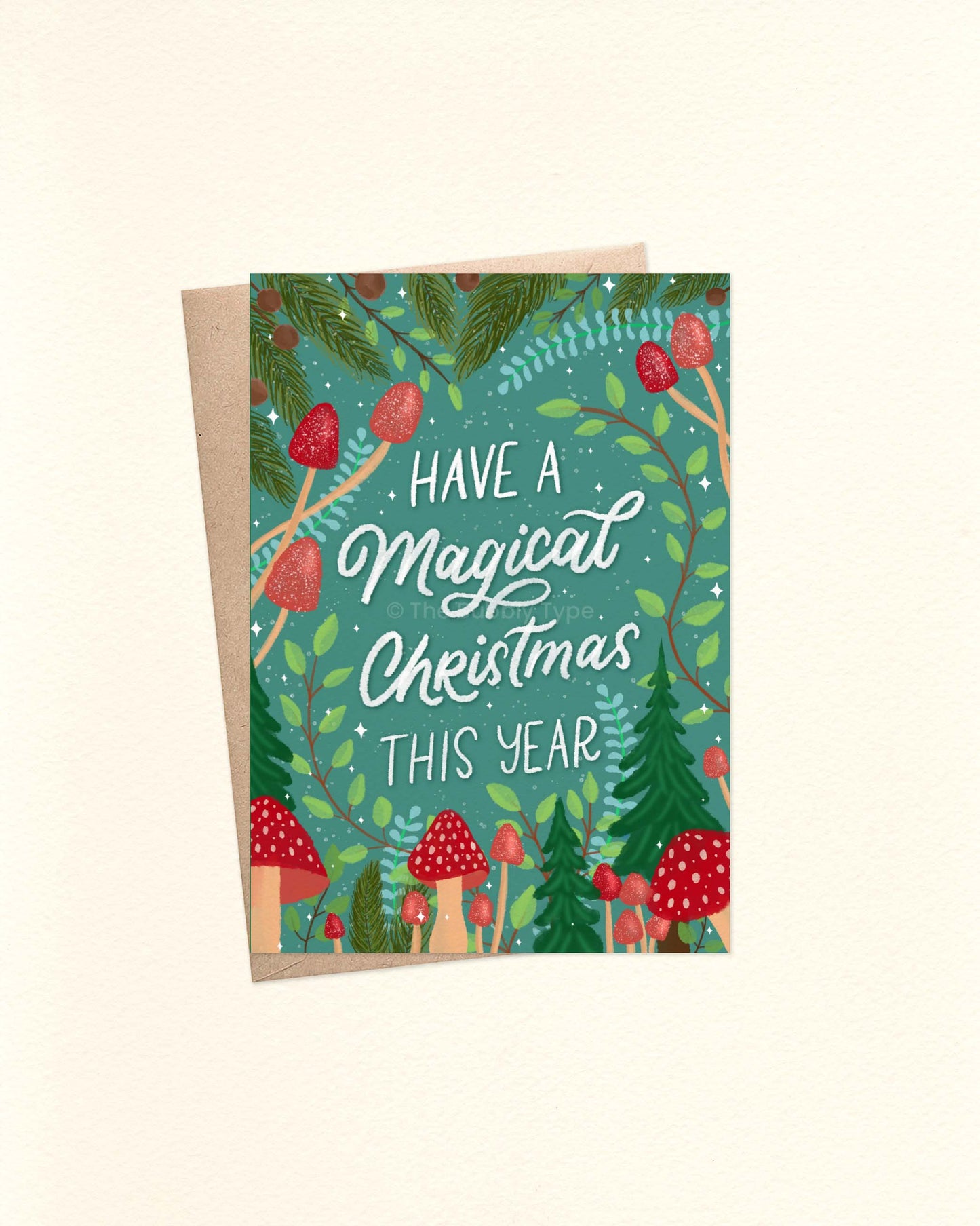 A6 Christmas card in teal green with red mushroom illustrations, pine tree foliage, and whimsical ‘Have a magical Christmas this year’ message, styled on a brown kraft envelope.