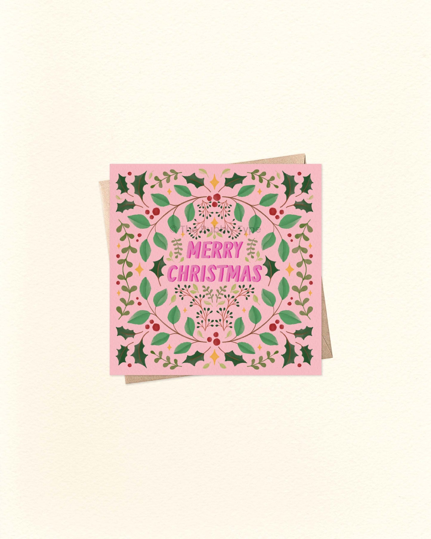 Square pink Christmas greeting card with festive foliage border and bold ‘Merry Christmas’ lettering in the centre, shown on a brown kraft envelope.