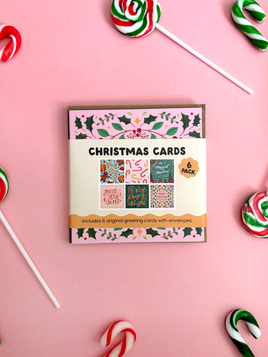 A boxed set of Christmas greeting cards laid flat on a pink background, surrounded by candy canes and lollipops. The package front reads 'CHRISTMAS CARDS' and '6 PACK' with small sample card designs visible.