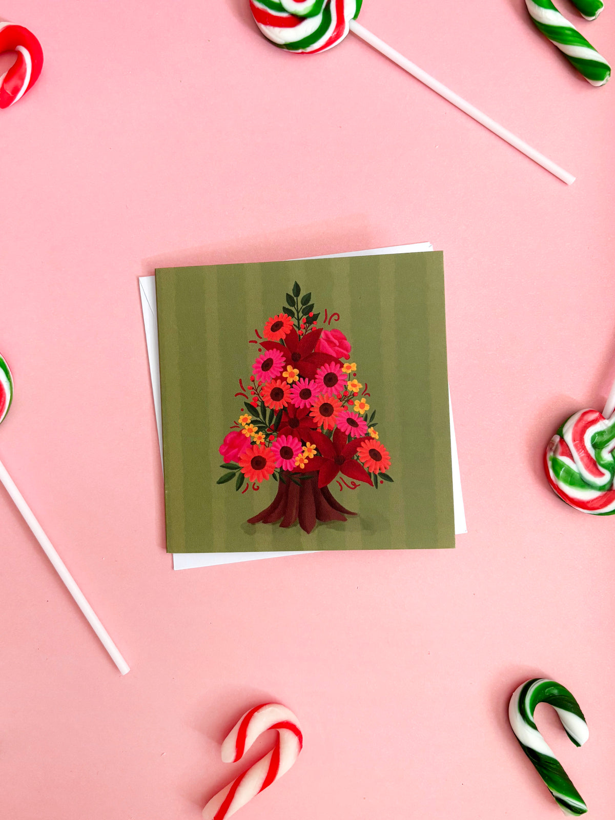 A square greeting card with a green striped background and an illustration of a Christmas tree made of colourful flowers, displayed on a pink background with candy cane props.
