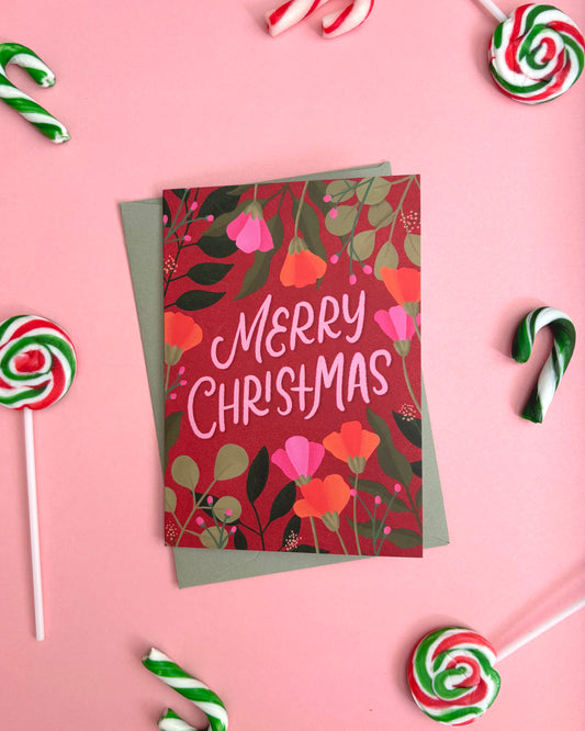 A Christmas greeting card with a red floral pattern and the words 'MERRY CHRISTMAS' in white script, layered over a muted green envelope. The card is photographed from above on a pink background, surrounded by candy canes and lollipops.