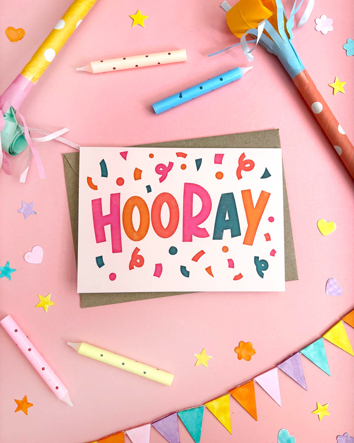 A greeting card featuring bold letters saying "HOORAY" with confetti surrounding it. The colourful design is laid on top of a brown kraft envelope on a pink paper background and surrounded by candles, party poppers, confetti and bunting flags.