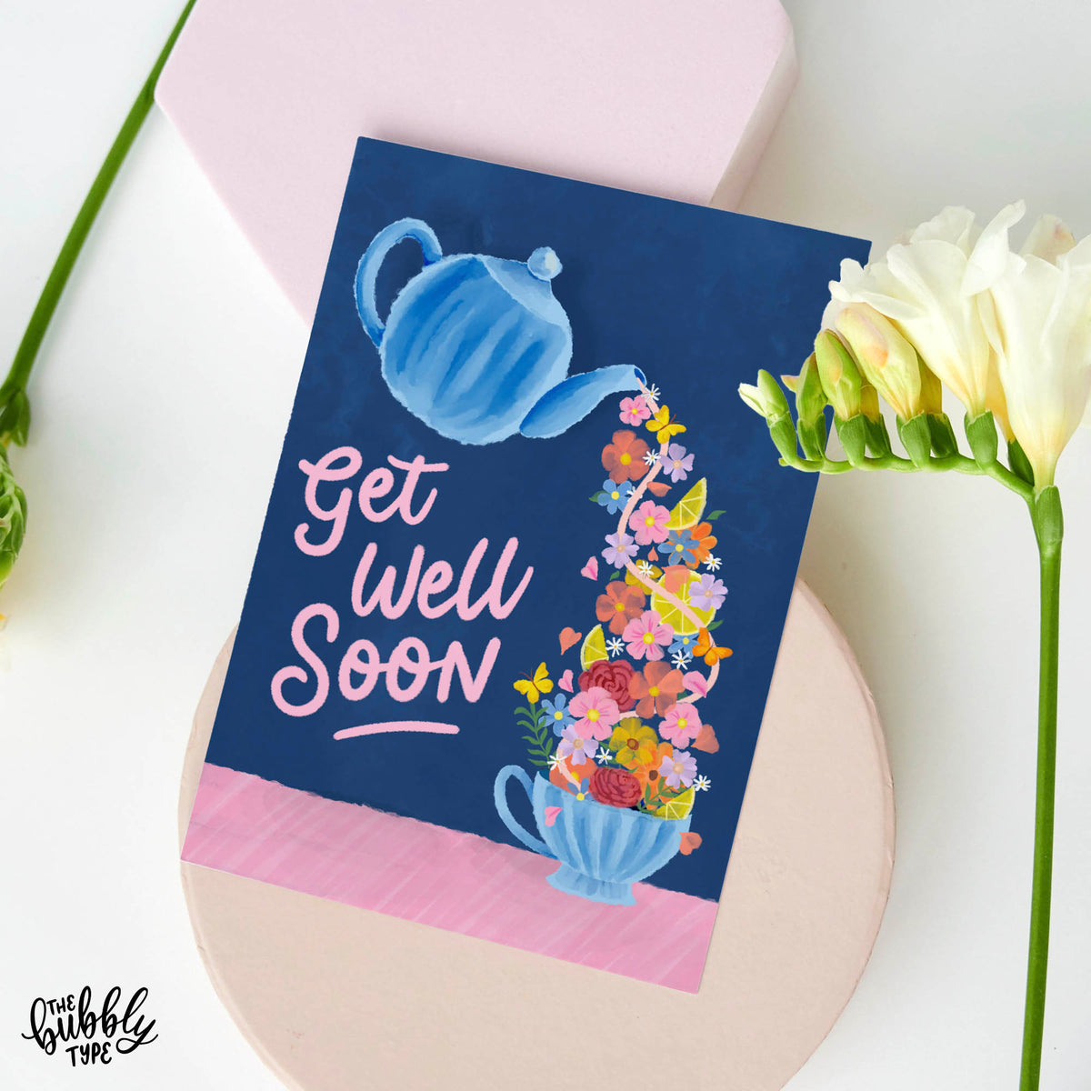 Greeting card with a watercolour-style blue teapot illustration tilted and pouring out a flow of yellow lemons, pink and orange flowers, green foliage, and butterflies into a matching blue teacup. The hand-lettered script reads “get well soon.”