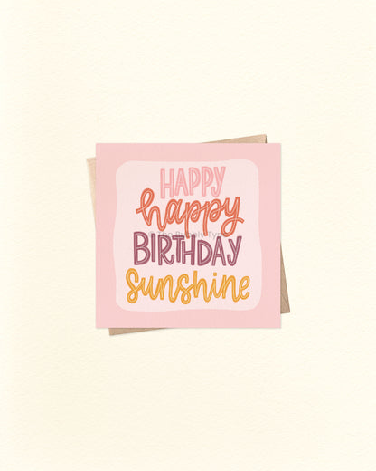 Front of 10cm square birthday card featuring the phrase “Happy Happy Birthday Sunshine” in vibrant watercolour lettering, with shades of purple, deep red, and yellow on a soft pink-purple background. Paired with a brown kraft envelope. A joyful, Australian made card by The Bubbly Type – perfect for brightening someone’s special day.