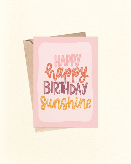 Front of A6 birthday card with a hand-lettered message that reads “happy happy birthday sunshine” in a loopy cursive font. The text features warm colours including pink, orange, warm purple, and yellow, set against a pale pinky-purple background with a rich purple border. A cheerful and uplifting birthday card from The Bubbly Type, shown with a brown kraft envelope.