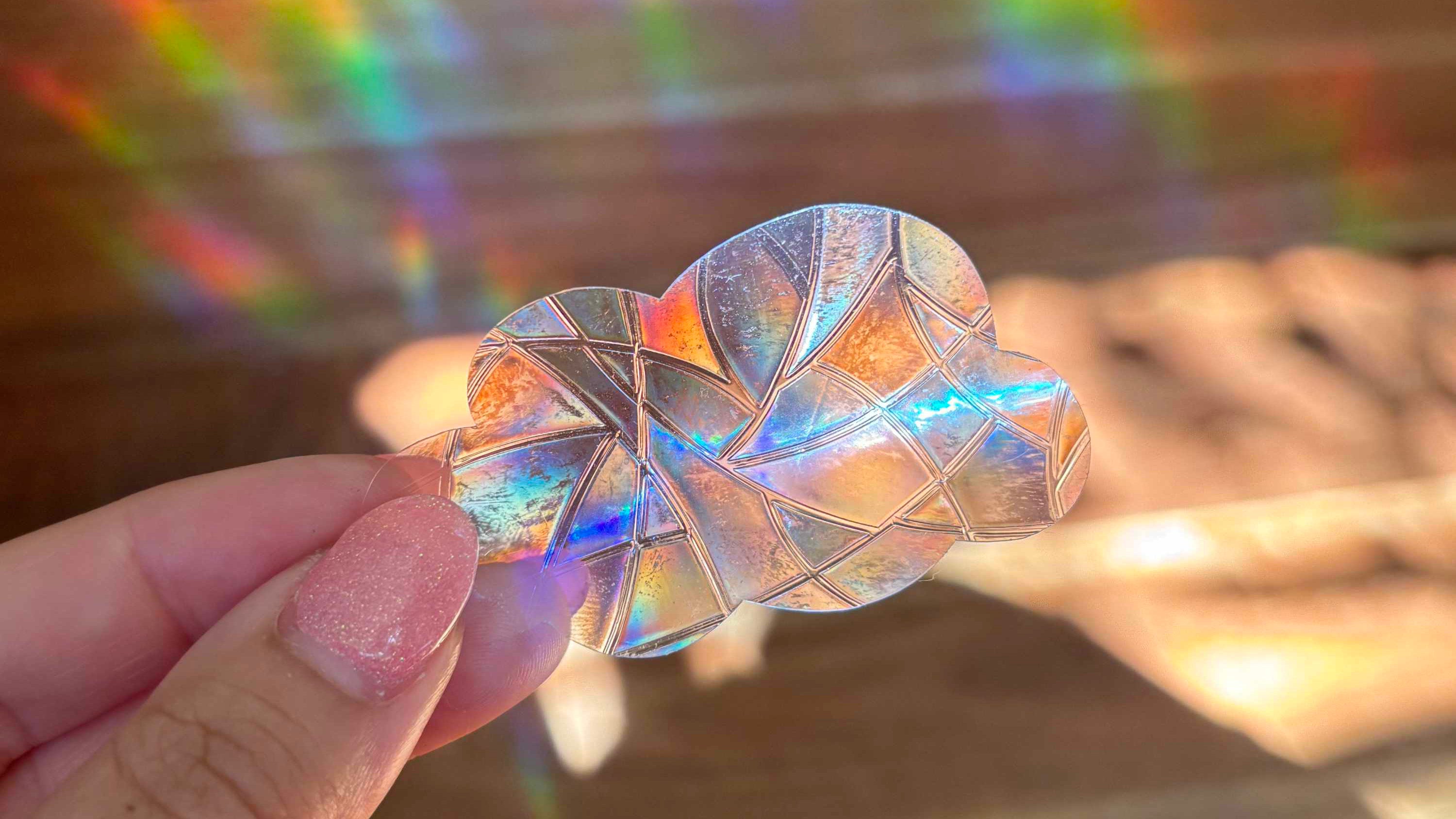 Iridescent sun catcher window sticker shaped like a cloud and held by a hand with a blurred, rainbow light in the background