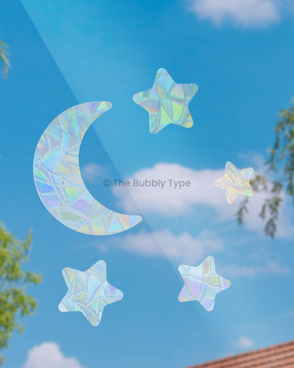 One crescent moon and four star-shaped sun catchers stuck on a window, magical space-themed rainbow maker decals, Australian made celestial window stickers for dreamy light reflections.