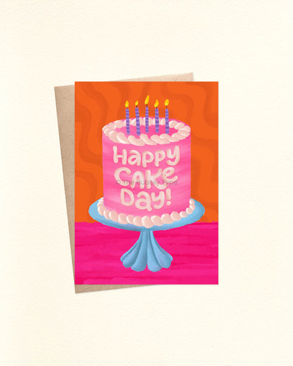 Front of an A6 birthday card featuring a watercolor illustration of a pink cake with pale pink-to-white frosting, sitting on a blue glass cake stand. The cake is topped with purple striped candles lit with yellow flames. The background is a vibrant hot pink and orange with a groovy wavy texture. The card is laid over a brown craft envelope.
