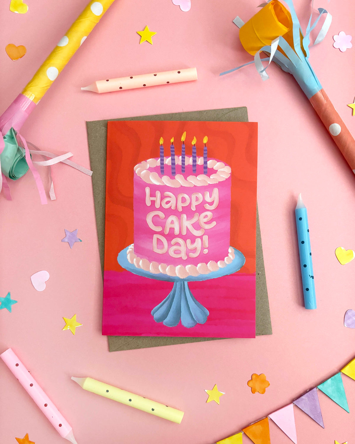 A greeting card featuring a pink birthday cake with candles and the words 'Happy CAKE Day!' in white, styled on a pink background with party props like candles, streamers, and star confetti.