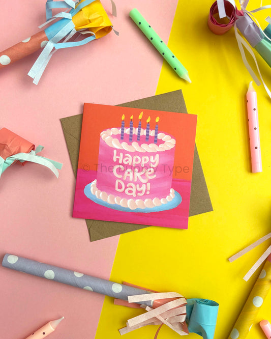 A square birthday card featuring a pink cake with candles and the words 'Happy CAKE Day!' in white, styled on top of a brown envelope with party supplies including coloured candles and paper party horns with streamers on a split pink and yellow background.