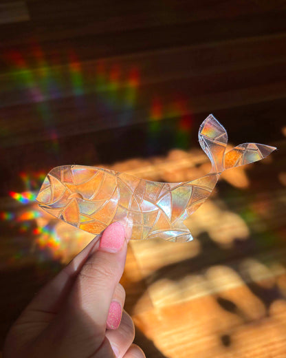 Iridescent whale-shaped sun catcher sticker held by a hand with pink nail polish, against a blurred background of rainbow light rays.