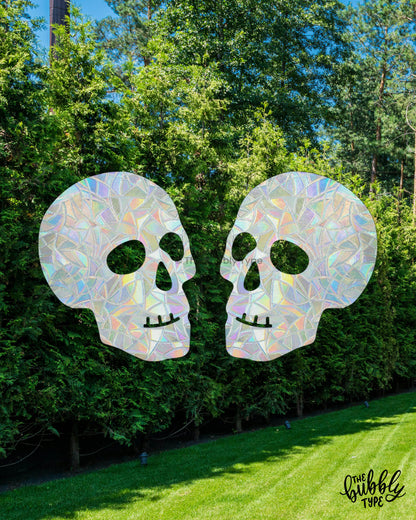 A pair of skull-shaped sun catchers facing each other on a window that overlooks a bright backyard with lush green grass, tall hedge trees, and a sunny blue sky. Perfect decor for Halloween or for witchy girls to enjoy all year round.