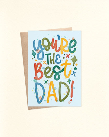 Front of A6 Father’s Day card with hand-lettered message: “You’re the best dad” in playful primary colours (red, blue, yellow and green) on a pale blue background. The design features watercolour splotches and splashes for a fun, artsy, craft-style vibe. Displayed with a brown kraft envelope.