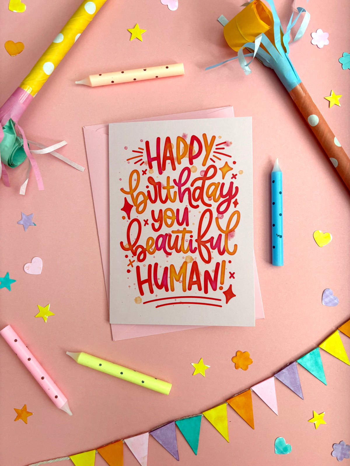 A birthday greeting card with colourful hand-lettered text reading 'HAPPY birthday you beautiful HUMAN!' placed on a pink surface surrounded by party props, star confetti, candles, and bunting.