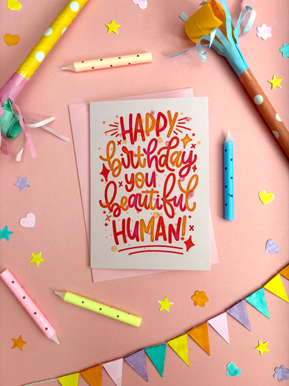 A birthday greeting card with colourful hand-lettered text reading 'HAPPY birthday you beautiful HUMAN!' placed on a pink surface surrounded by party props, star confetti, candles, and bunting.
