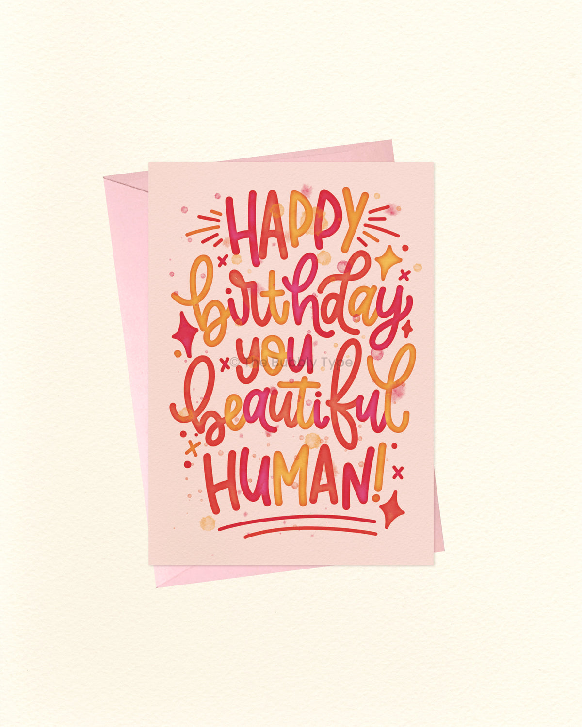 A greeting card with a pink envelope behind it, featuring colourful hand-lettered text that reads “HAPPY birthday you beautiful HUMAN!” on a warm off-white background.