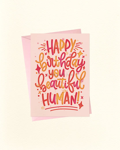 A greeting card with a pink envelope behind it, featuring colourful hand-lettered text that reads “HAPPY birthday you beautiful HUMAN!” on a warm off-white background.