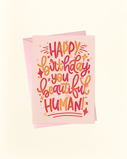 A greeting card with a pink envelope behind it, featuring colourful hand-lettered text that reads “HAPPY birthday you beautiful HUMAN!” on a warm off-white background.