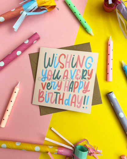 A square birthday greeting card with colourful hand-lettered text reading 'Wishing you a very, very Happy Birthday!!' placed on top of a brown envelope. The card is photographed in a styled flat-lay with pastel party supplies, including rolled paper, party hats, and candles, arranged on a split pink and yellow background.