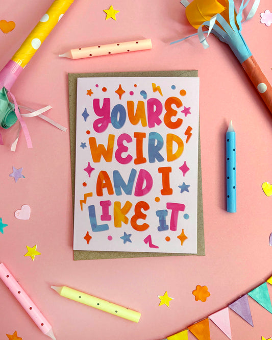 A greeting card with colourful retro-style lettering that reads 'YOU'RE WEIRD AND I LIKE IT' on a white cardstock with a brown kraft envelope, styled on a pink background with party candles and confetti.