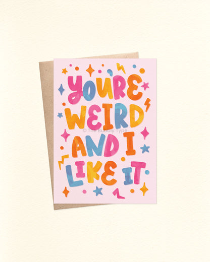 Front of A6 greeting card with playful block hand-lettering that reads: “You’re weird and I like it.” The text appears in bright watercolour-style cutout letters in pink, blue, yellow, and orange on a pale pink background. Surrounded by colourful shape accents including stars, lightning bolts, dots, and squares. Shown with a brown kraft envelope.