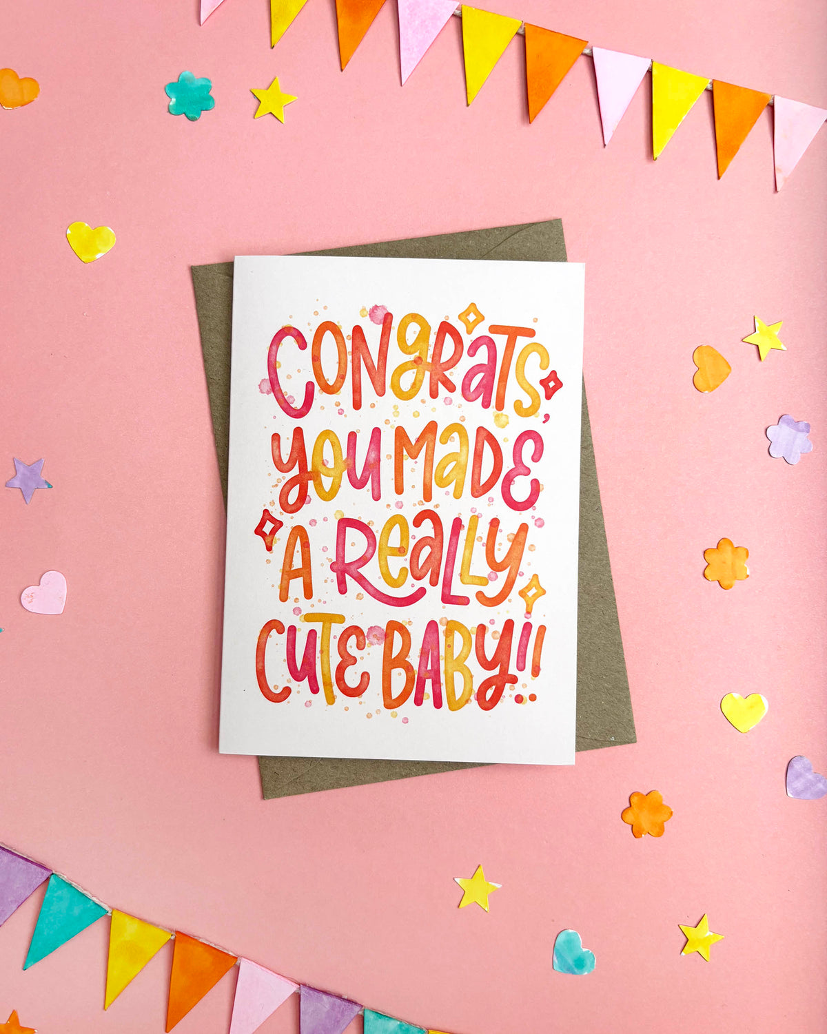 A white greeting card with colourful hand-lettered text reading 'CONGRATS YOU MADE A REALLY CUTE BABY!!' placed on top of a brown envelope, photographed on a pink background with bunting, stars, hearts, and flowers.