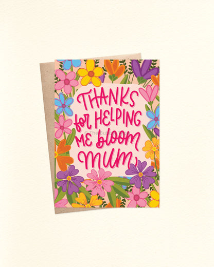 Front of A6 Mother’s Day card featuring a bright floral border in solid colours and line art style. Flowers in yellow, orange, purple, pink, and blue surround the hand-lettered message: “Thanks for helping me bloom mum.” Shown with a brown kraft envelope.