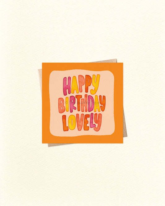 Front of 10cm square birthday card featuring hand-lettered watercolour text that reads “Happy Birthday Lovely” in the centre, surrounded by an orange border and lighter orange background. Paired with a brown kraft envelope. Australian made card by The Bubbly Type – a bright and cheerful birthday greeting for someone special.