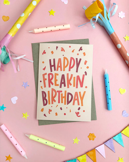 A birthday greeting card reading “HAPPY FREAKIN BIRTHDAY” in colourful hand-lettered text, placed on a pink surface with party props including candles, confetti, paper stars, and party hats.