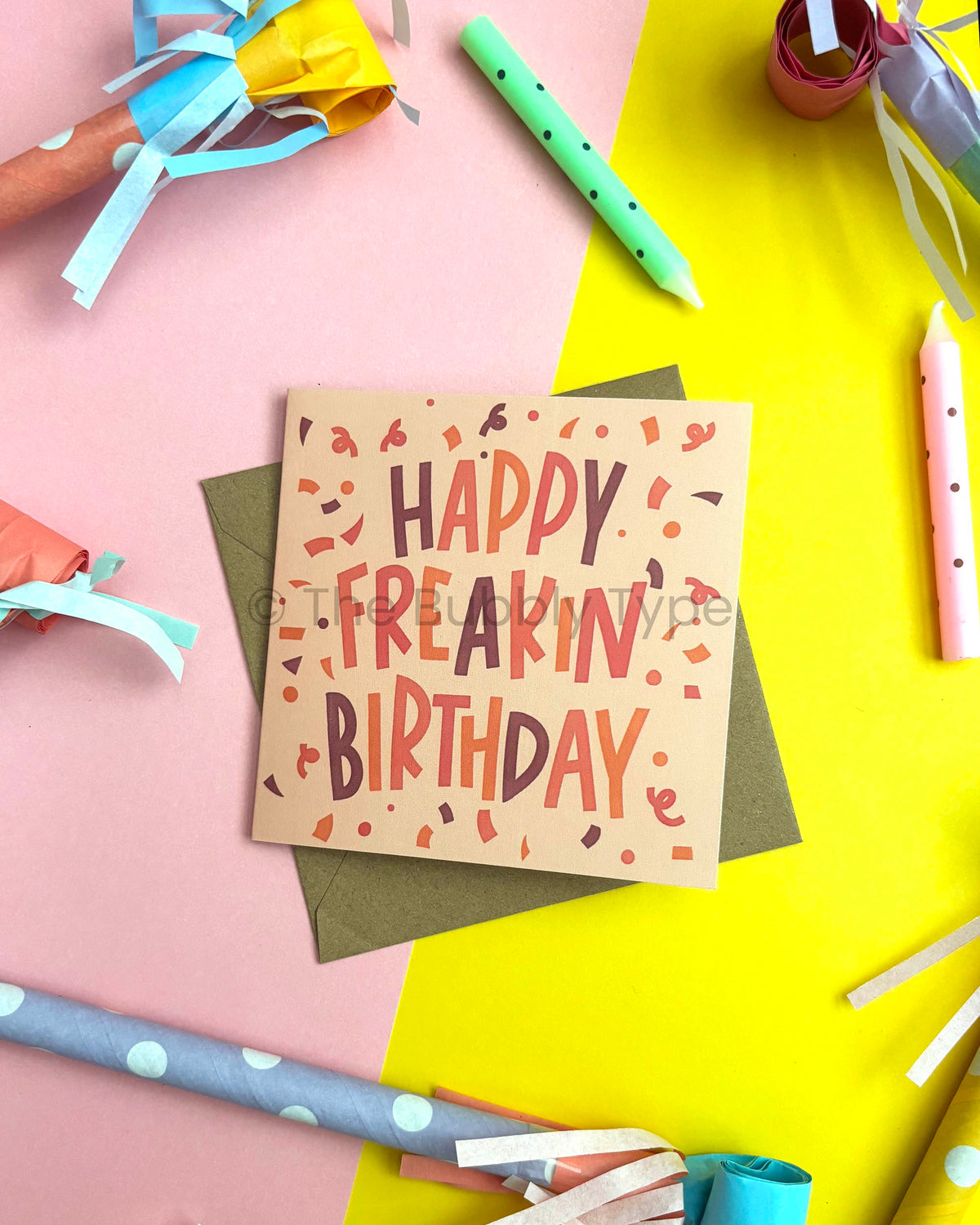 A square birthday card reading “HAPPY FREAKIN’ BIRTHDAY” in stylized lettering, placed on top of a kraft brown envelope. The card is photographed in a styled flat-lay with party supplies, confetti illustrations, and a pink and yellow background.