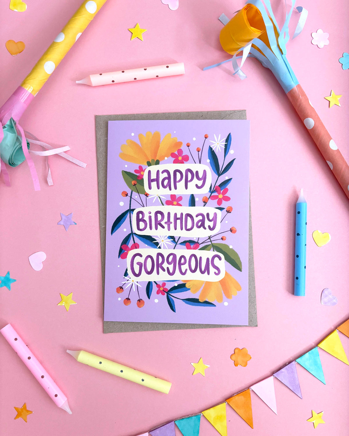 A birthday greeting card with a lavender background and colourful floral illustration, reading 'Happy Birthday Gorgeous' in white lettering. The card is placed on a pink surface surrounded by party props including candles, streamers, star confetti, and bunting.
