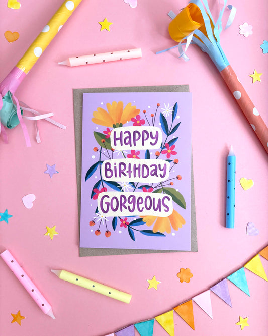 A birthday greeting card with a lavender background and colourful floral illustration, reading 'Happy Birthday Gorgeous' in white lettering. The card is placed on a pink surface surrounded by party props including candles, streamers, star confetti, and bunting.