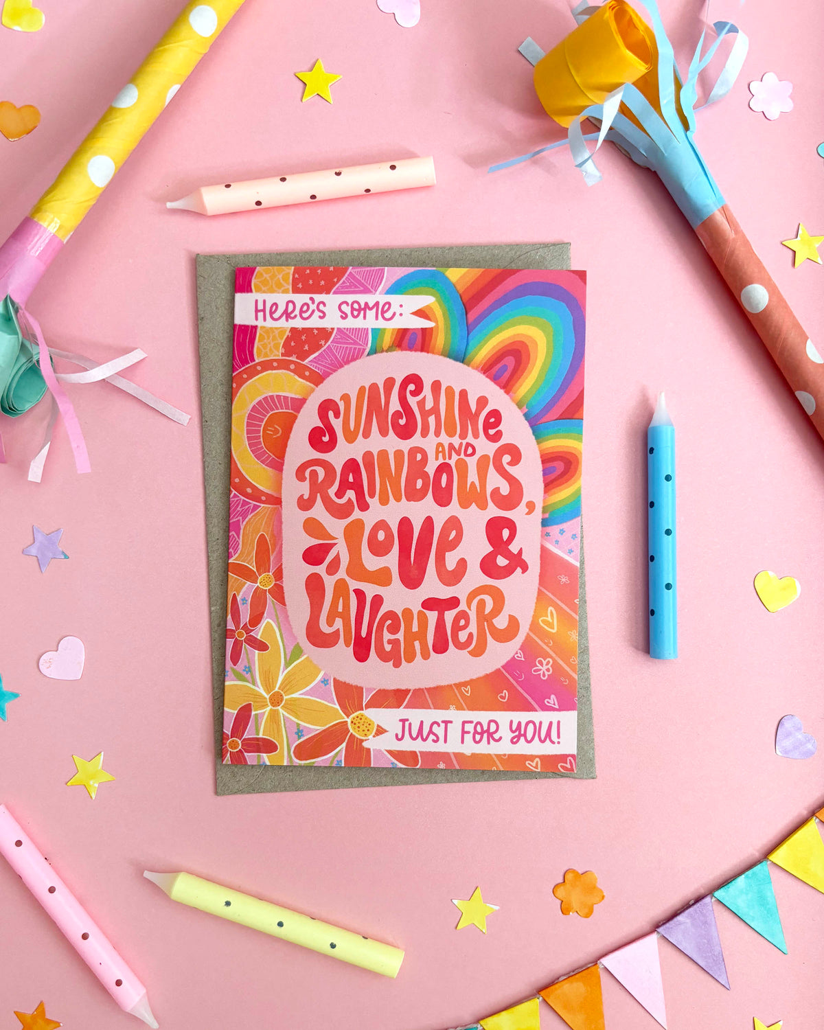 A colourful greeting card with retro rainbow swirls and the text 'Sunshine and Rainbows, Love & Laughter' is displayed on a pink background with party-themed props like candles, paper stars, and bunting.