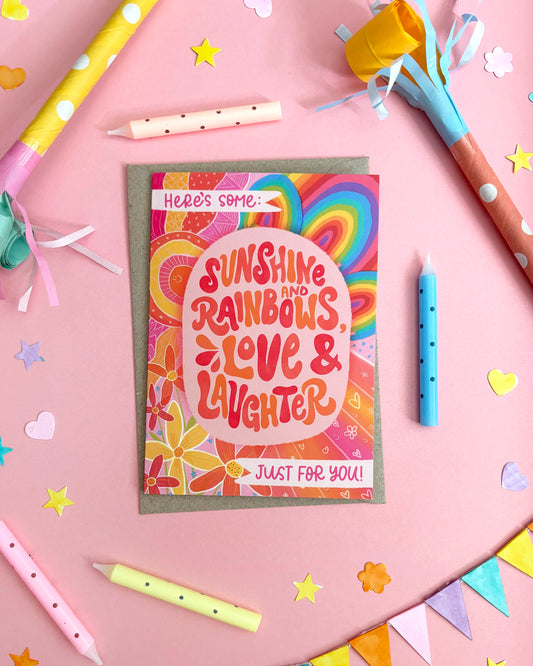A colourful greeting card with retro rainbow swirls and the text 'Sunshine and Rainbows, Love & Laughter' is displayed on a pink background with party-themed props like candles, paper stars, and bunting.