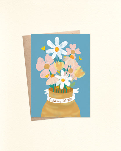 A6 greeting card with a dusty blue background. The illustration features a mustrad coloured vase filled with pink, white and yellow flowers, and green foliage in a watercolour style. A white ribbon wraps around the neck of the vase with hand-lettered text reading “Thinking of You.” Shown with a brown kraft paper envelope (included with the sale).