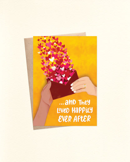 Front of A6 wedding card featuring a yellow watercolour background with a hand-drawn illustration of two different hands holding a red envelope. One hand wears a diamond engagement ring. A joyful burst of red love hearts rises from the envelope. At the bottom, bold white hand-lettered text reads: “... and they lived happily ever after.” Shown with a brown kraft envelope. Use for weddings, engagements, bridal shower and more.