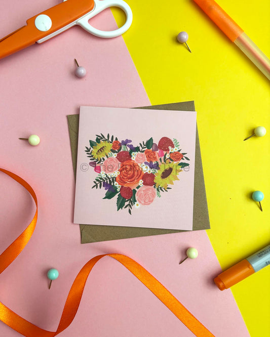 A square greeting card with a colourful floral heart design, placed on top of a brown envelope. The card is photographed in a styled flat-lay arrangement on a split pink and yellow background with orange scissors, push pins, and an orange marker visible around it.
