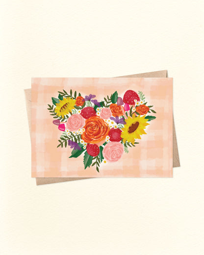 Front of A6 landscape greeting card featuring a whimsical heart-shaped cluster of colourful flowers, mushrooms, and foliage. The background is a soft peach watercolour gingham pattern. Perfect for weddings, anniversaries, Mother's Day, or everyday love notes. Displayed with a brown kraft envelope.