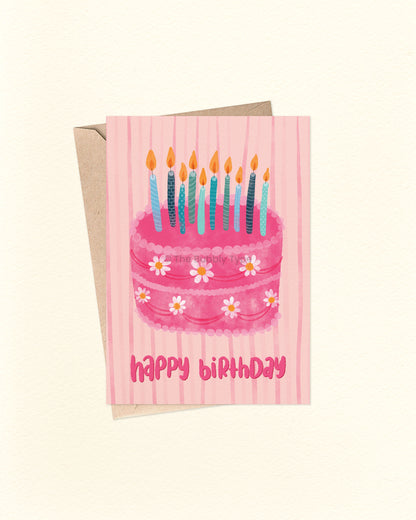 Front of A6 birthday card featuring a whimsical illustration of a pink cake decorated with white daisy icing flowers and topped with a cluster of blue and teal candles. Hand-lettered “Happy Birthday” sits below the cake on a pale pink background with soft watercolour line textures. Shown with a brown kraft envelope.