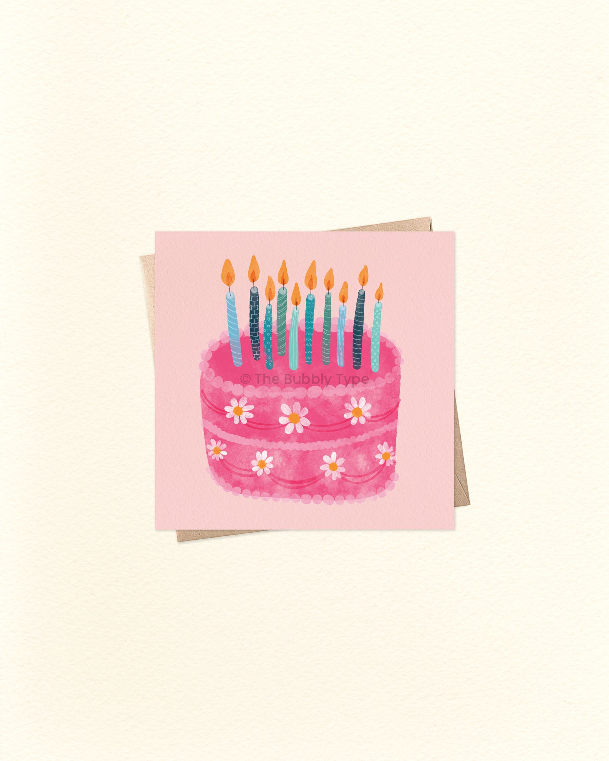 Front of 10cm square birthday card with a watercolour illustration of a pink birthday cake decorated with white daisy flowers and topped with multiple blue and teal candles. Set on a soft pink background and paired with a brown kraft envelope. A sweet and whimsical Australian made card by The Bubbly Type – perfect for birthdays of all ages.