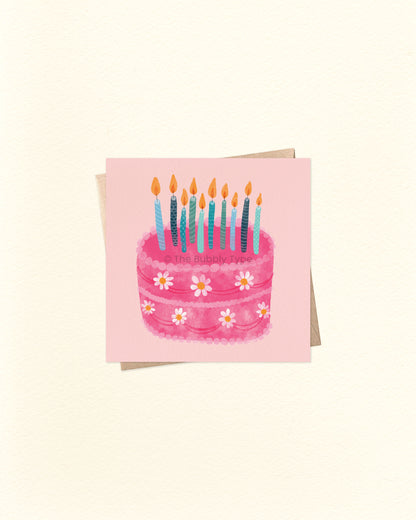 Front of 10cm square birthday card with a watercolour illustration of a pink birthday cake decorated with white daisy flowers and topped with multiple blue and teal candles. Set on a soft pink background and paired with a brown kraft envelope. A sweet and whimsical Australian made card by The Bubbly Type – perfect for birthdays of all ages.