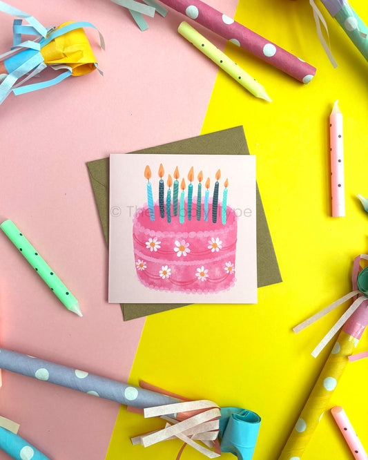 A square greeting card featuring a watercolour-style pink birthday cake with blue candles and small white flowers, placed on a Kraft envelope. The card is photographed in a styled flat-lay arrangement with colourful paper rolls, ribbons, and candles on a split pink and yellow background.