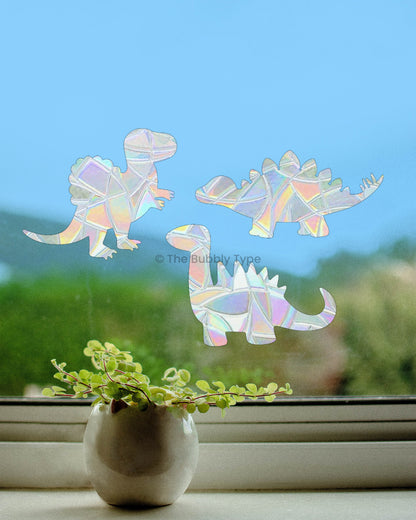 Dinosaur-shaped rainbow making suncatcher stickers facing each other, stuck to a window with a dramatic blue sky and greenery as the backdrop—creating a peaceful, whimsical atmosphere. there is little pot plant on the window sill in the foreground.